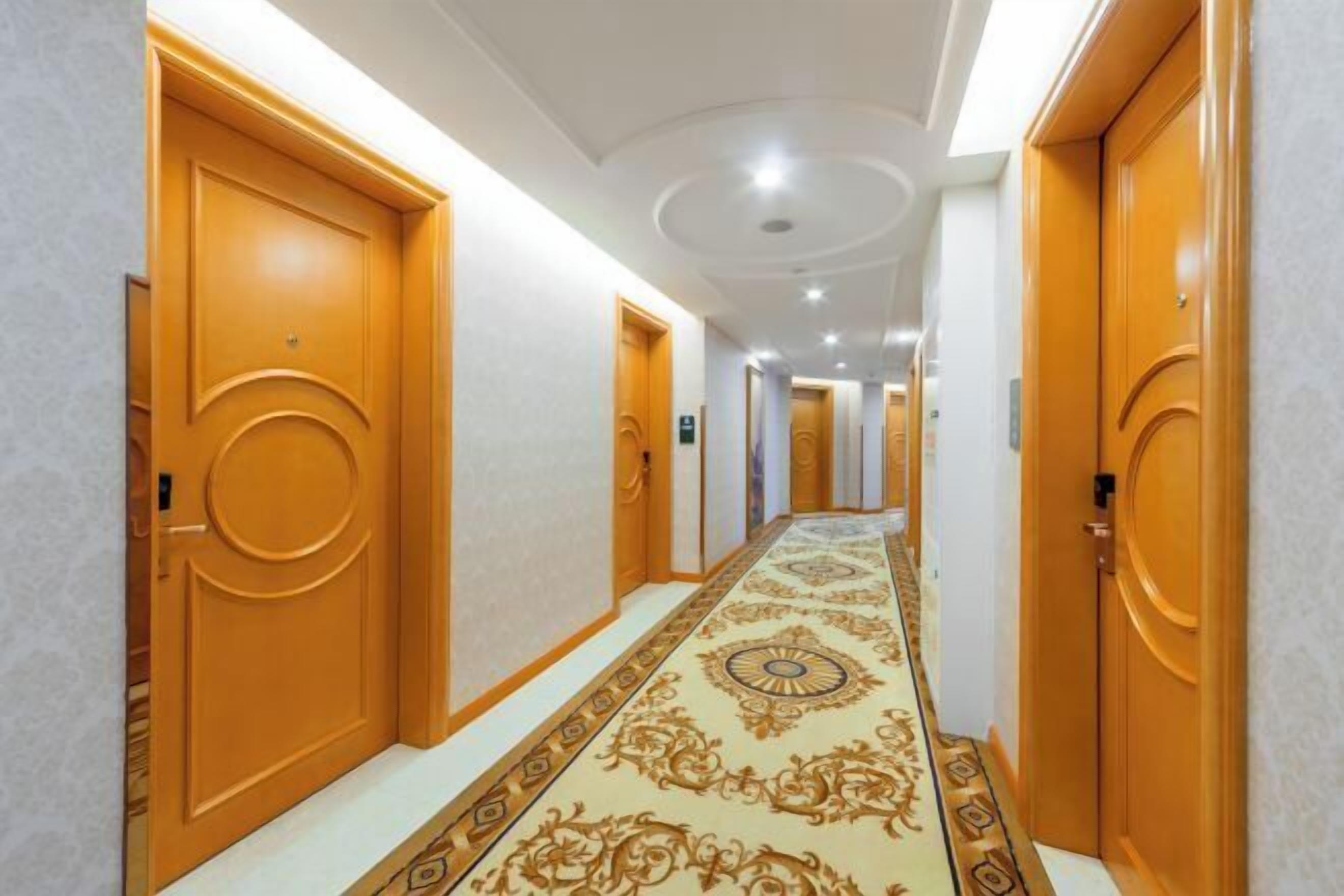 A hotel hallway with ornate carpet, wooden doors, and a modern ceiling design.