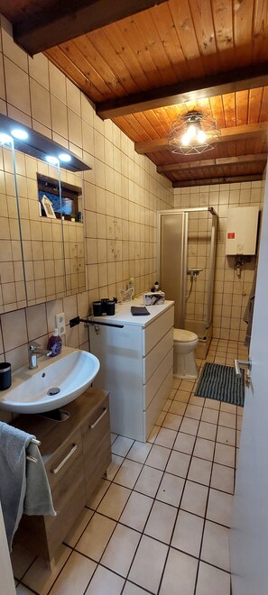 A bathroom with a wooden ceiling, tiled walls, a white sink, a toilet, and a shower area. - Ferienwohnung im Schönen Odenwald (Mörlenbach)