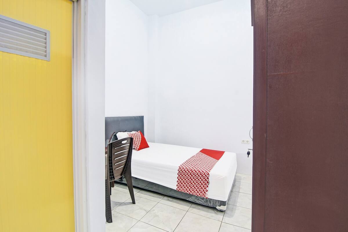 A small, single-bed room with a white bedspread and red accents, a wooden chair, and a yellow wall.