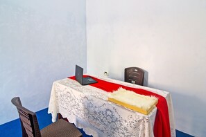 Une pièce avec une table recouverte d’une nappe en dentelle blanche et d’un tissu rouge, un ordinateur portable posé sur la table et une chaise avec un coussin foncé.