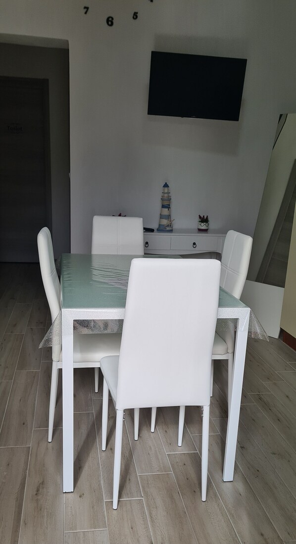 A dining area with a table and four chairs, a television mounted on the wall, and a small decorative item on the table. - Renovated 2-room apartment, parking space, by the sea. (Marina di Caronia)
