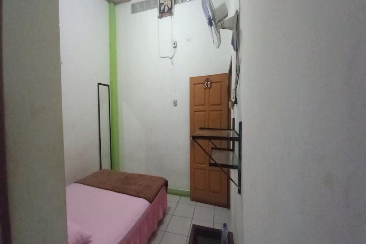 A small room with a bed, a door, a fan, and a shelf.