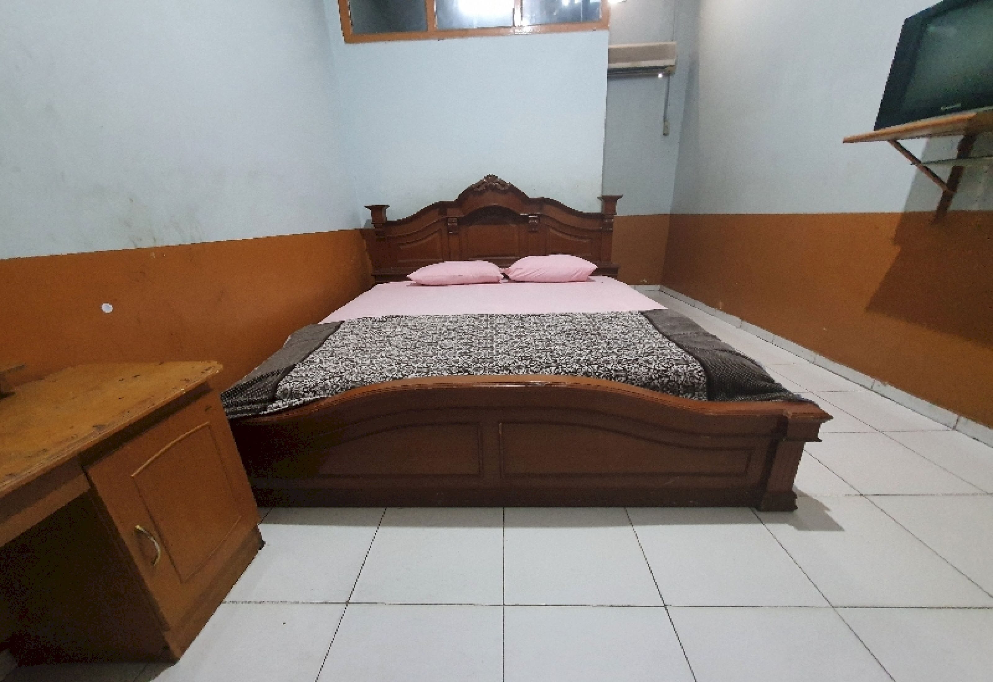 A bedroom with a wooden bed, a bedside table, a TV on a stand, and a window.