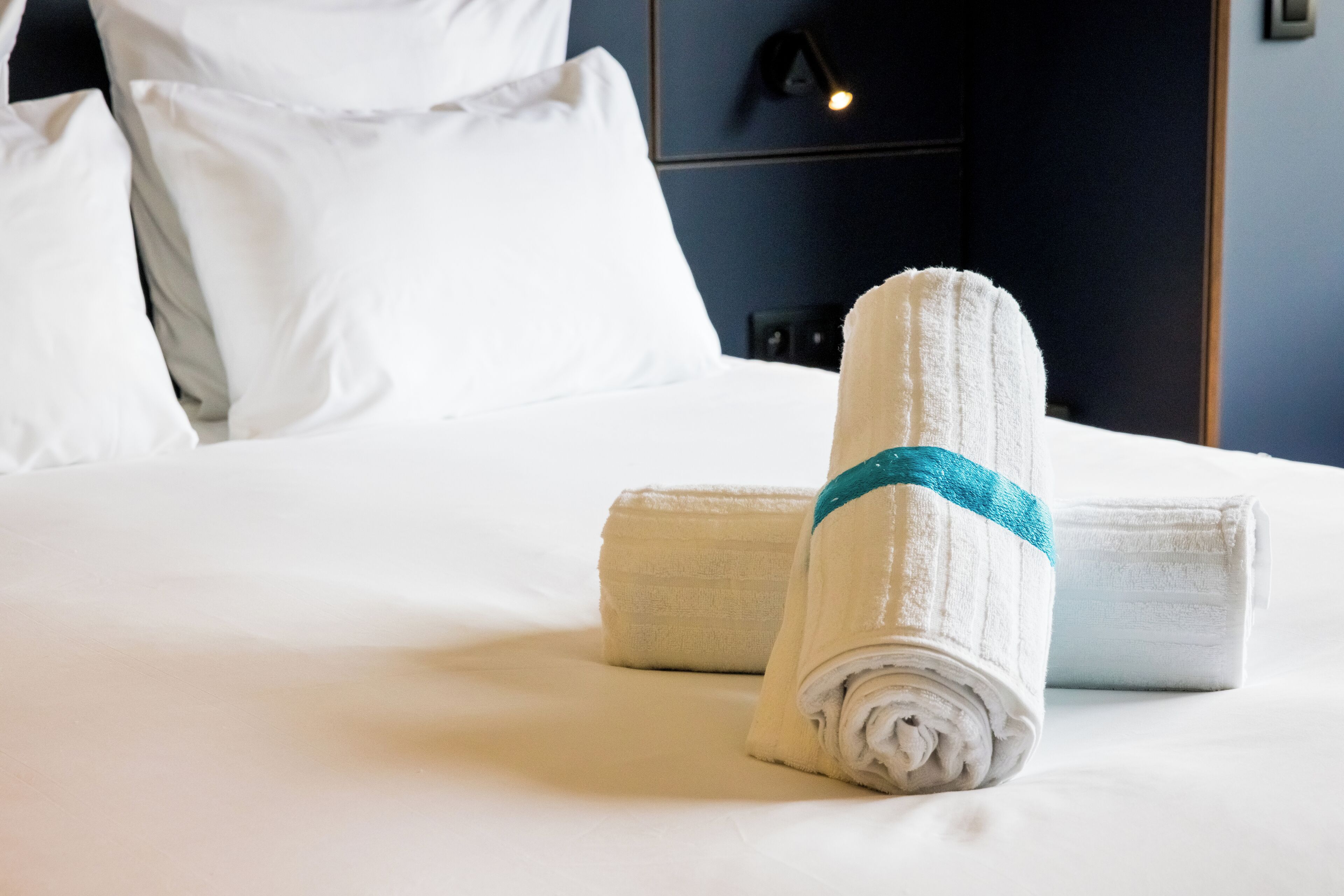 Una cama bien hecha con sábanas blancas y una toalla enrollada con un detalle azul.