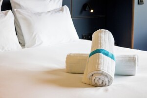 Una cama bien hecha con sábanas blancas y una toalla enrollada con un detalle azul.