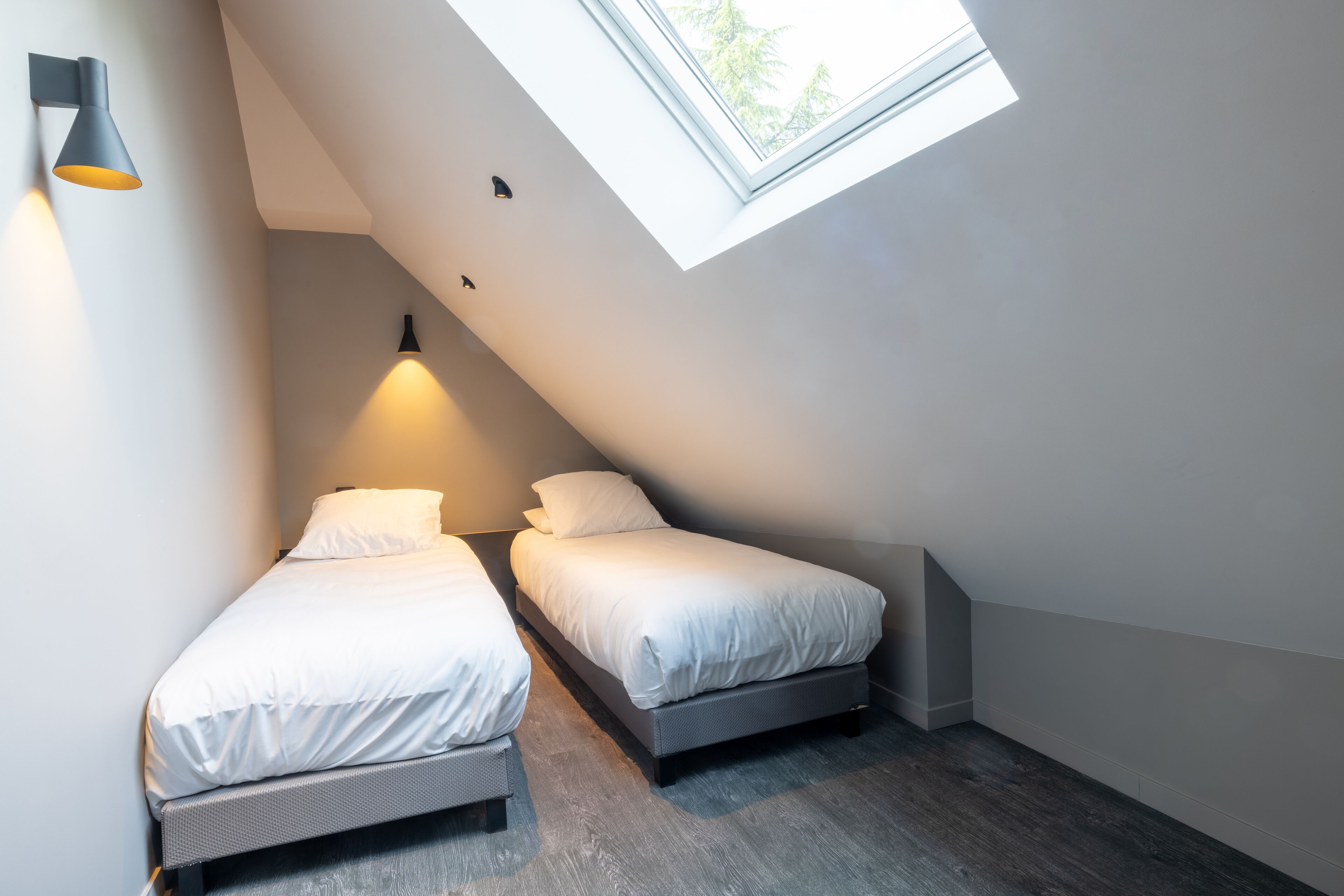 A small, modern bedroom with two single beds, a skylight, and a wall-mounted light fixture.