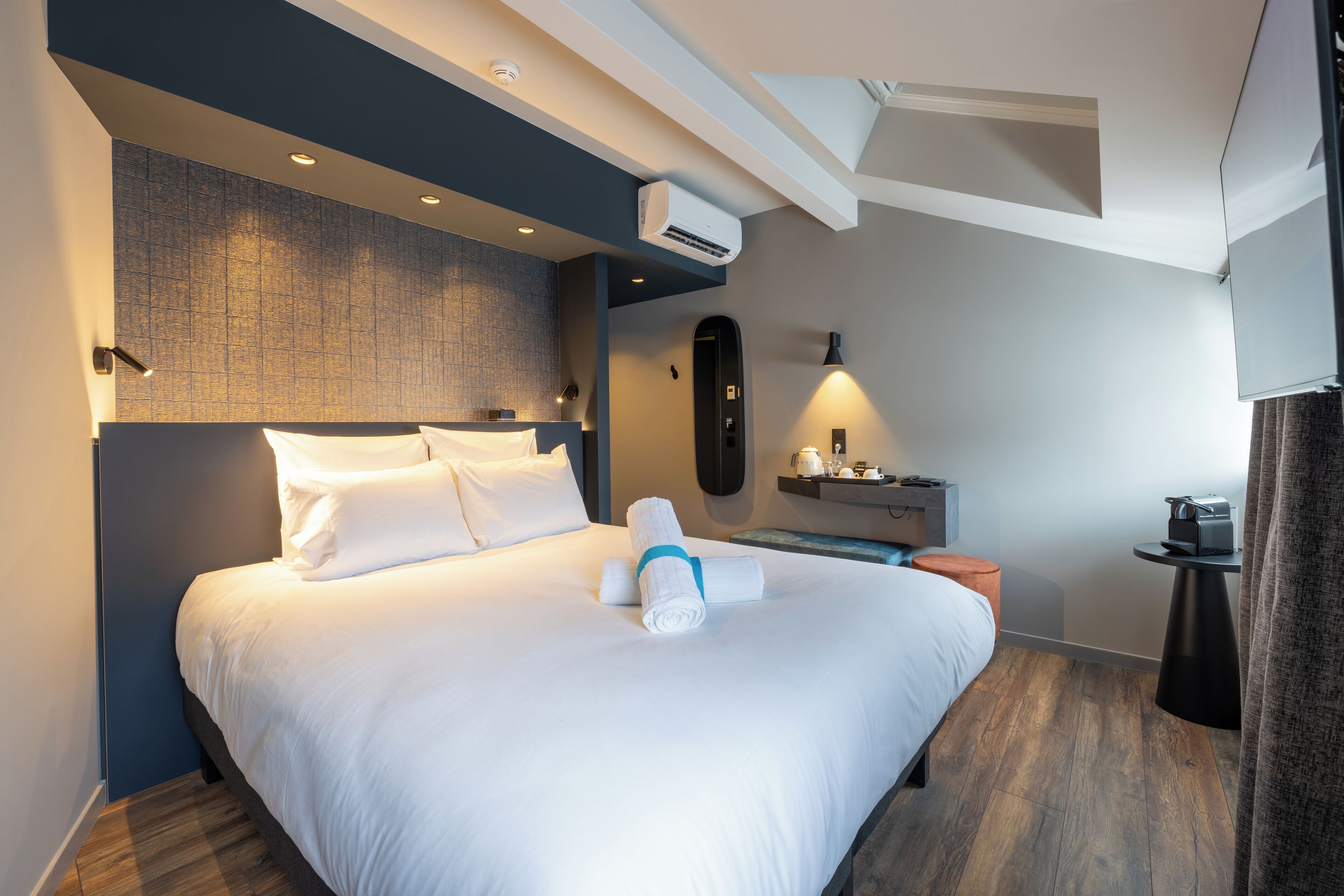 A modern hotel room with a neatly made bed, an air conditioning unit, a small desk with a lamp, and a coffee machine.