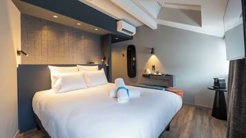 A modern hotel room with a neatly made bed, an air conditioning unit, a small desk with a lamp, and a coffee machine.