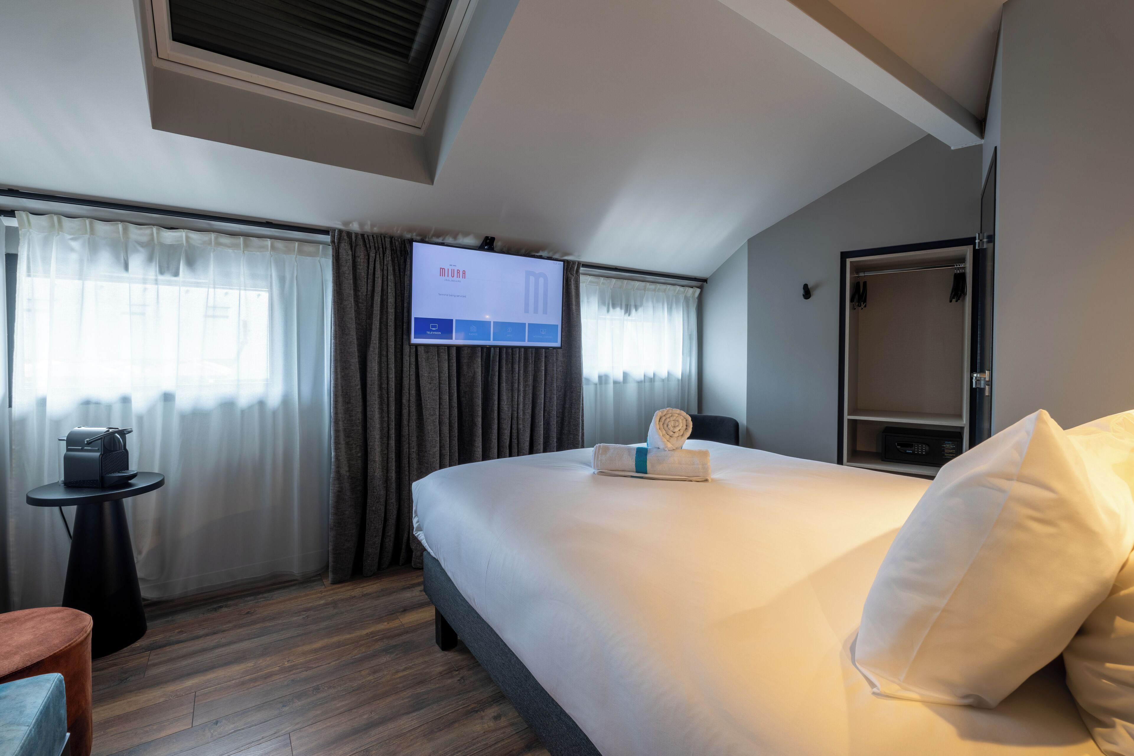 Habitación de hotel con una cama grande, un televisor de pantalla plana en la pared, una mesita con una cafetera y una puerta a la derecha.