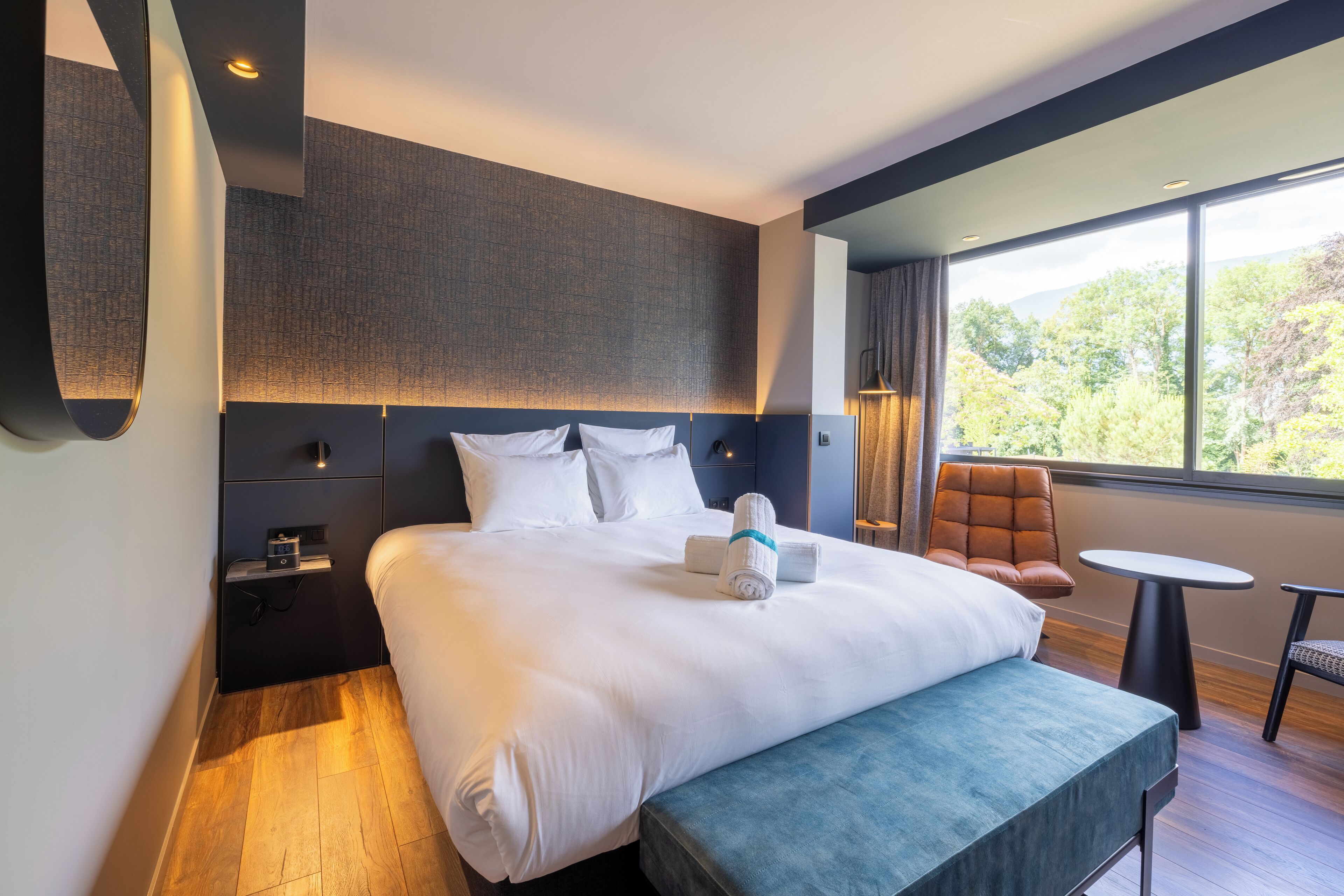 A modern hotel room with a large bed, a bench, a small table, and a view of the outdoors.