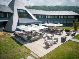 Um pátio ao ar livre com várias mesas e cadeiras, cercado por guarda-sóis, e um edifício moderno com design de telhado único ao fundo.