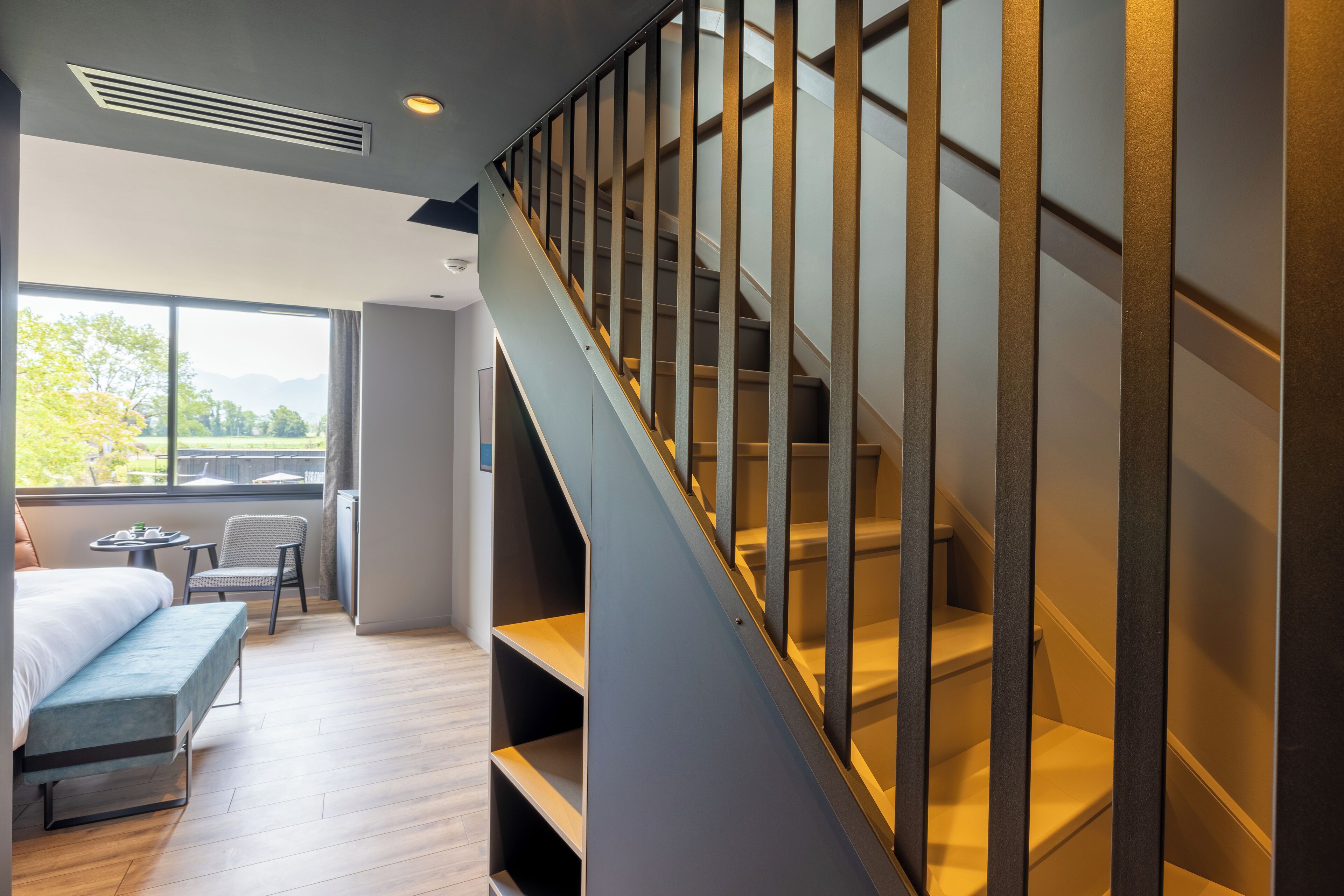 A modern hotel room with a staircase, a bed, a seating area, and a view of the outdoors.