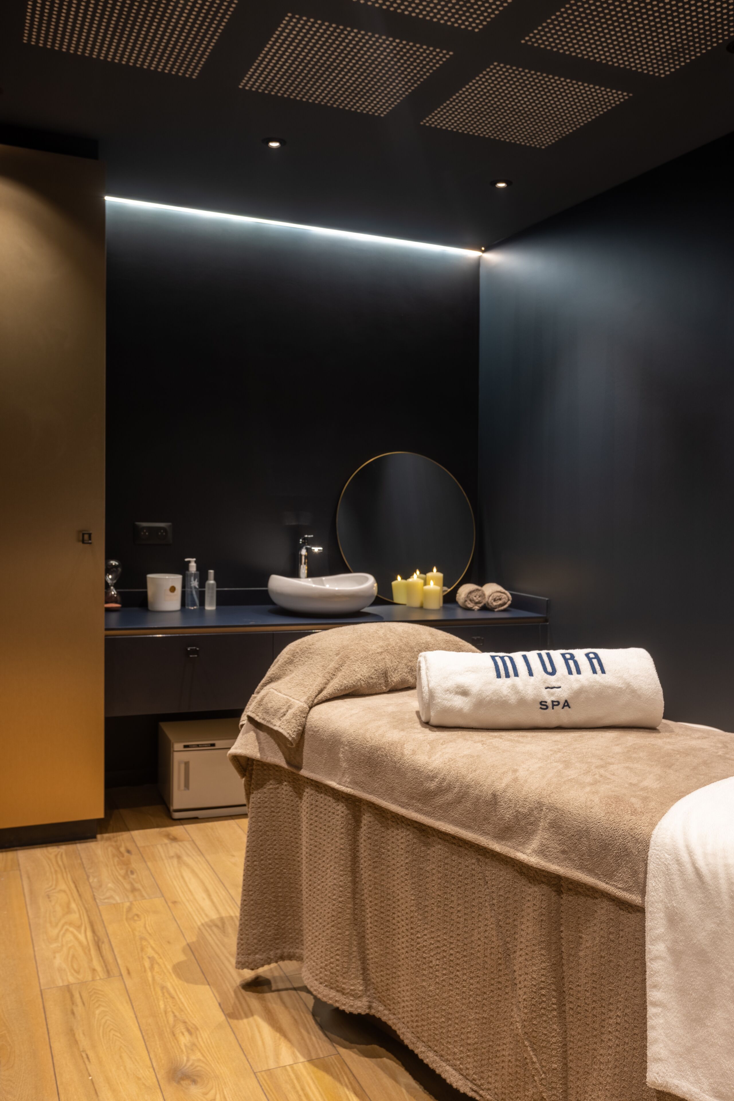 A modern spa room with a large bed, a vanity with a mirror, and a ceiling with a geometric pattern.