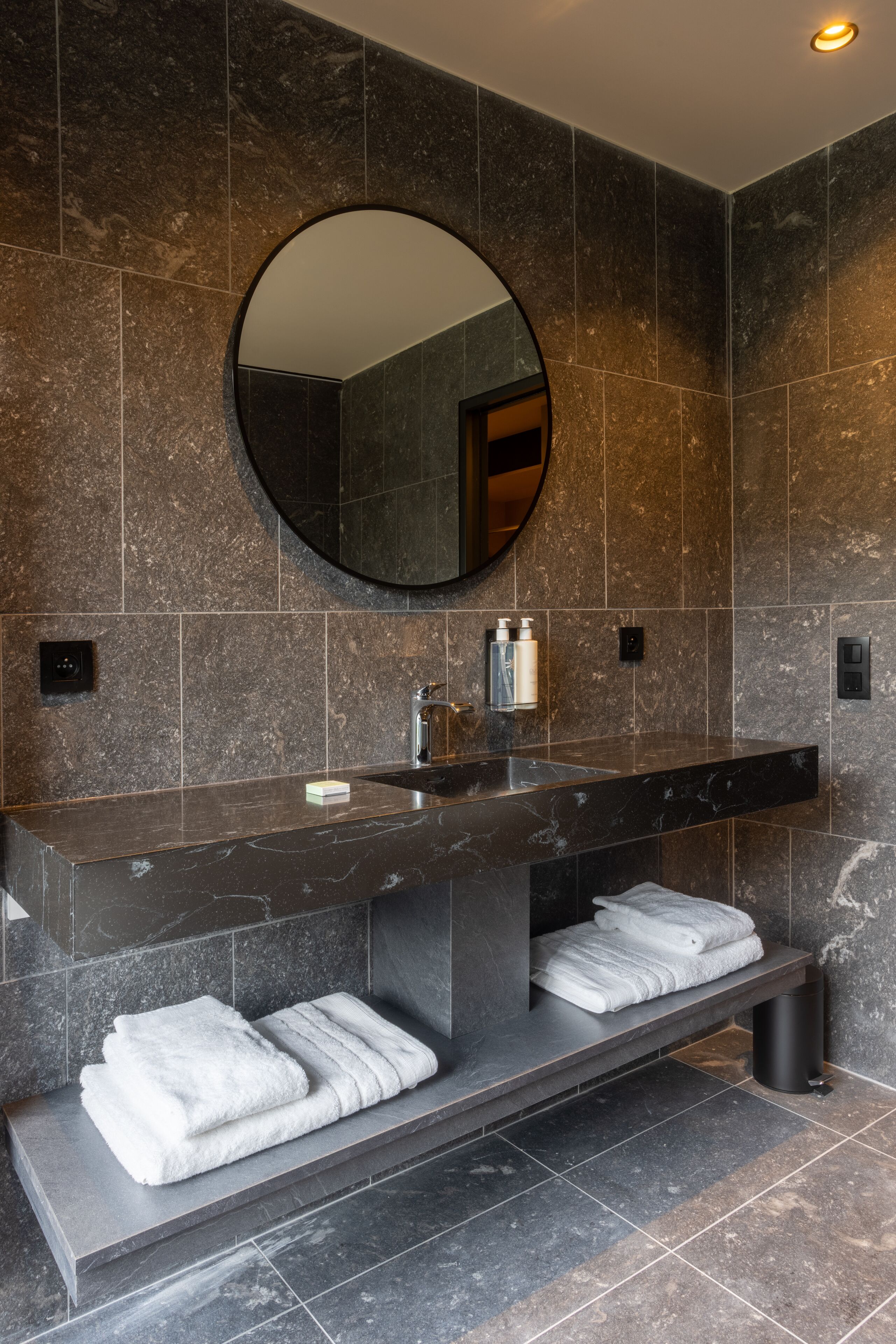 Un baño moderno con un lavamanos de mármol oscuro, un espejo grande y toallas ordenadas en un estante.