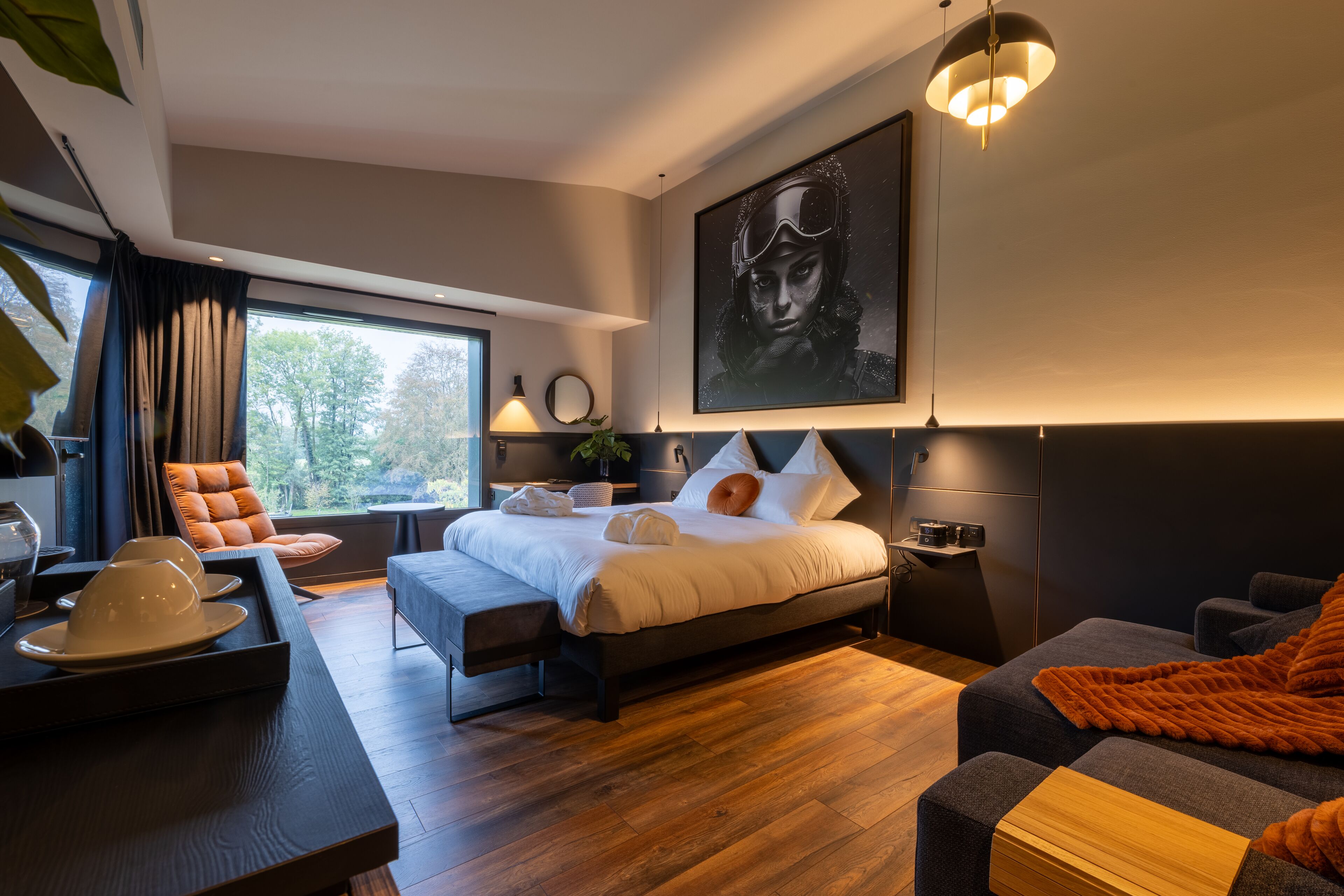 A modern bedroom with a large bed, a seating area with a sofa and armchair, a dining table with a tray, and a large window with curtains.