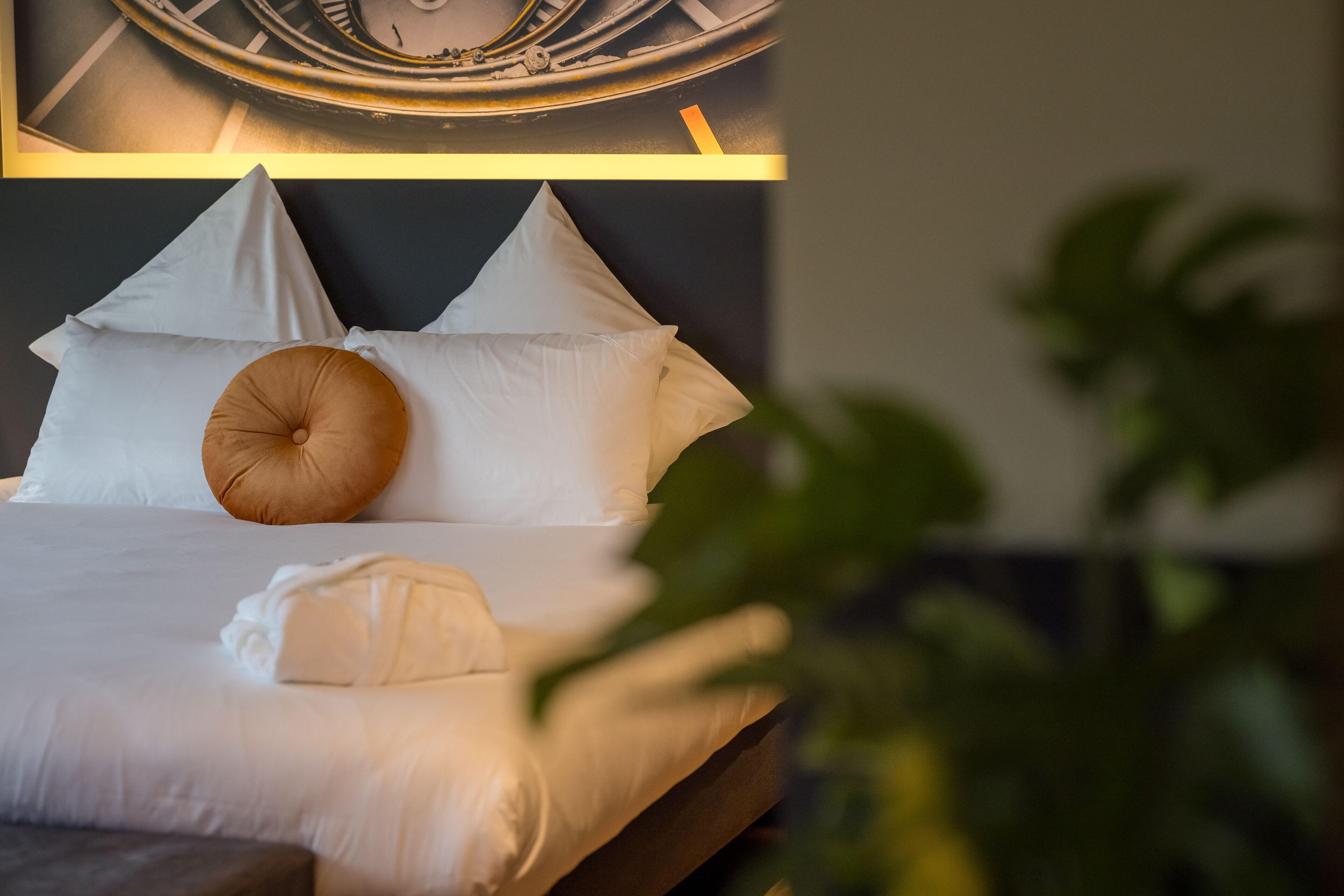 Una cama bien hecha con sábanas blancas y un cojín marrón decorativo, un reloj en la pared y una planta en primer plano.