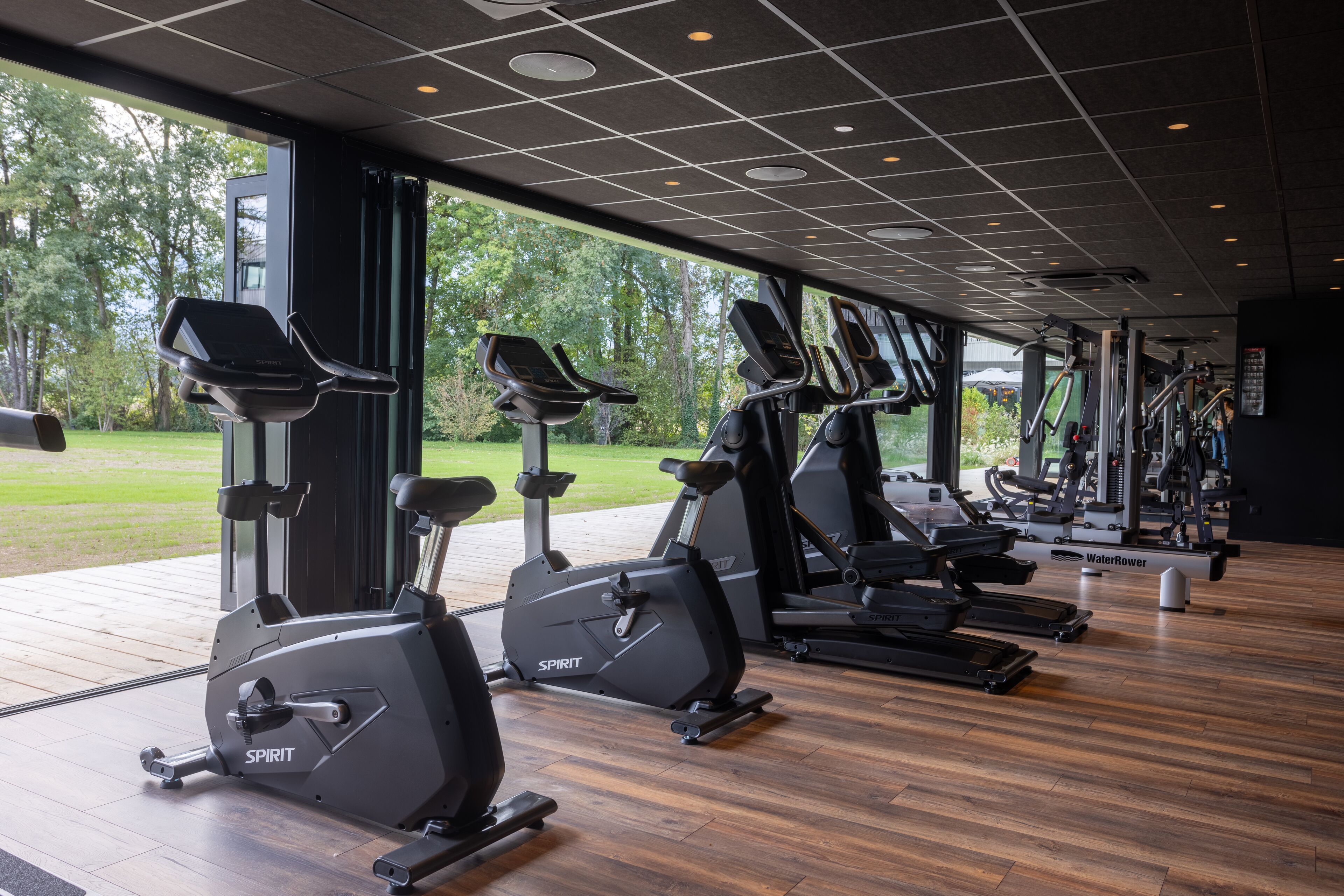 Un gimnasio con varias máquinas de cardio, ventanas amplias y suelo de madera.