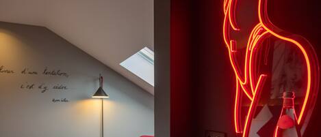A modern hotel room with a bed, a red neon flamingo sign, a bottle of water, and a small table with a lamp.