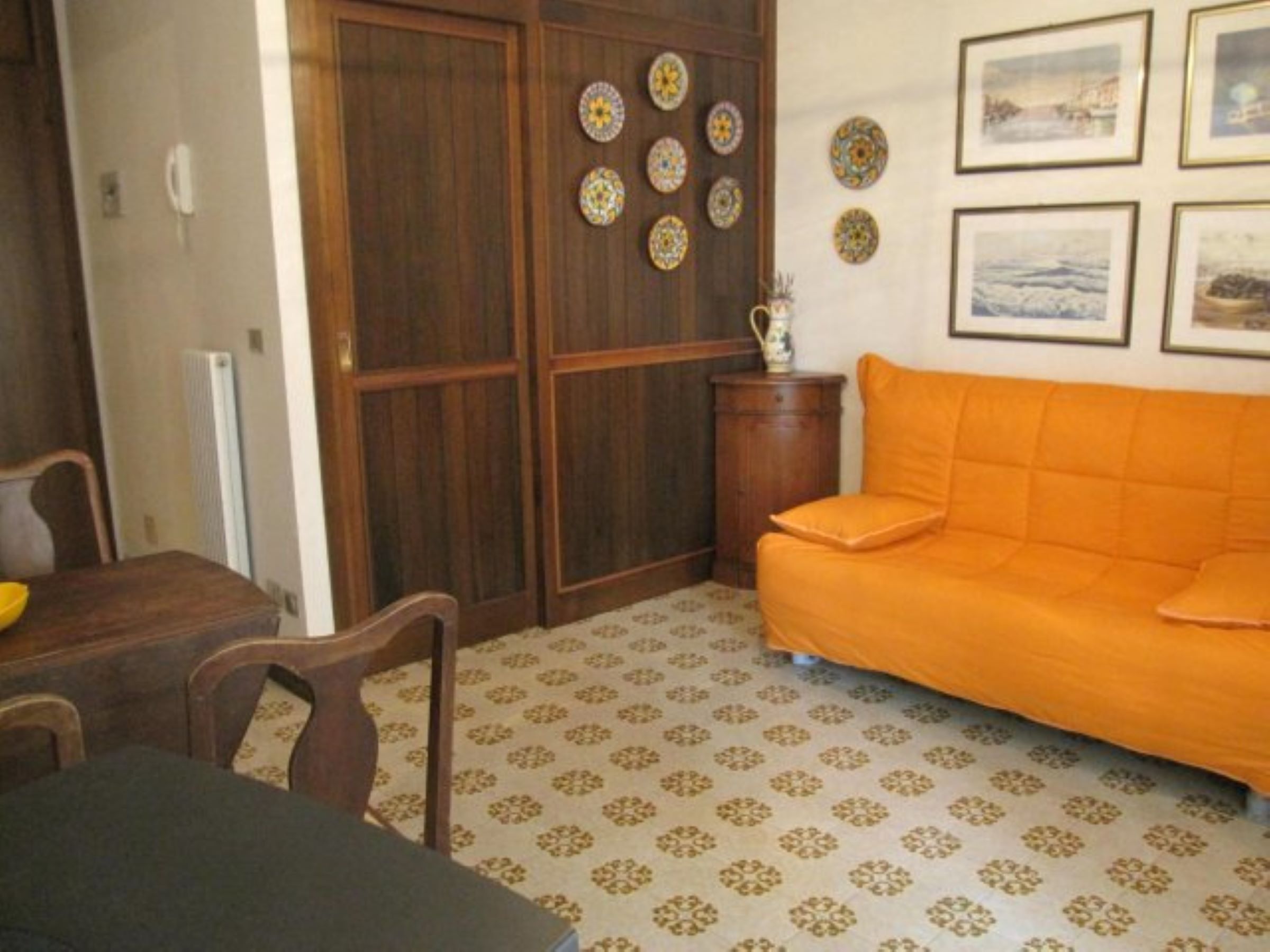 A room with a wooden wardrobe, a dining table with chairs, an orange sofa, patterned carpet, and framed pictures on the wall.