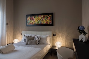 Quarto de hotel com cama, mesa de cabeceira com abajur, cadeira e um quadro pendurado na parede.