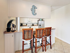 A kitchen with white cabinets, a microwave, a refrigerator, and a seahorse decoration on the wall. There are three wooden chairs with white ties at the kitchen island.