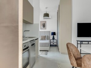 Ein modernes Hotelzimmer mit Küchenzeile, einem Bett mit Kopfteil, einem Nachttisch mit Lampe und einem Flachbildfernseher.