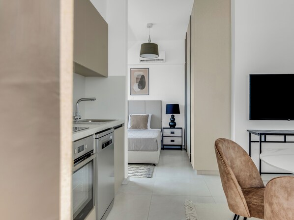 A modern hotel room with a kitchenette, a bed with a headboard, a bedside table with a lamp, and a flat-screen TV. - Sanders Elgio Limassol - Full Building (Limassol)