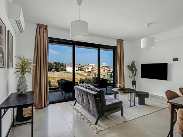 Un salon moderne avec un canapé, une table à manger et un téléviseur. Il y a une grande fenêtre avec des rideaux, une plante et une vue sur les bâtiments à l’extérieur.