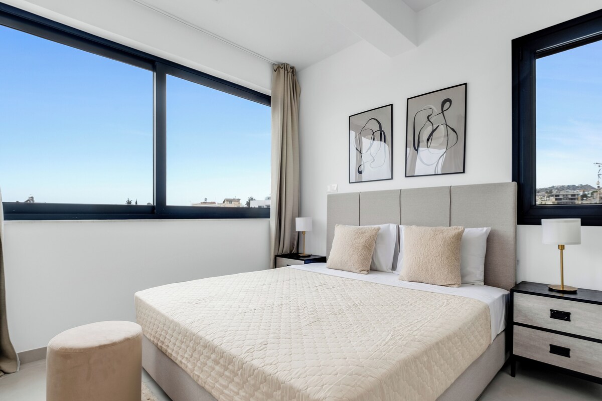A modern bedroom with a large bed, a nightstand, a round ottoman, and two wall-mounted lamps.