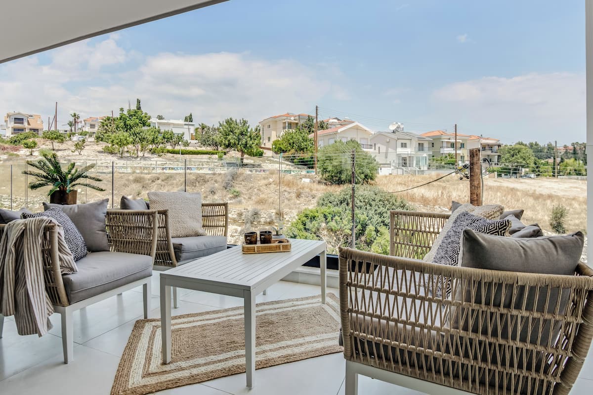 Un balcón con mobiliario de mimbre, una mesa blanca y vistas a una zona residencial con casas y vegetación.