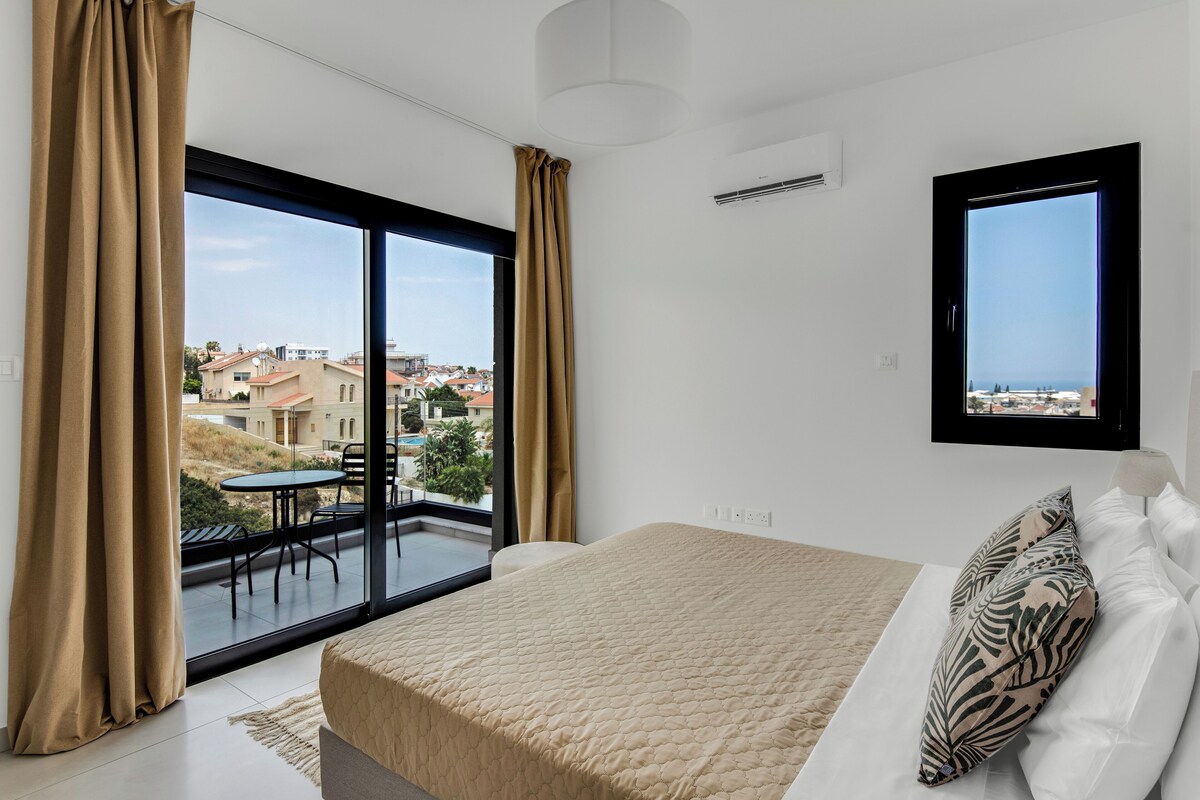 A bedroom with a large bed, a view of the city through a balcony, and a ceiling fan.
