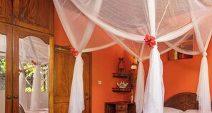 A room with a bed under a white canopy, a wooden door, and a wall-mounted shelf with decorative items. - Room-effies Mountain View Villa - Gecko (Mahé Island)