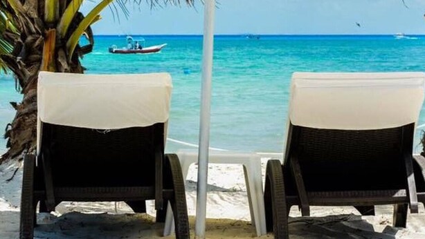 Deux fauteuils de plage en osier, dotés de coussins blancs, sur une plage de sable, avec des palmiers et un bateau en arrière-plan.