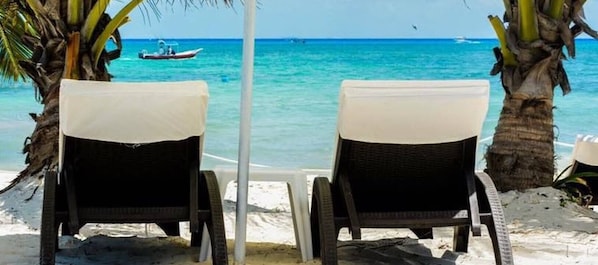 Dos sillones de mimbre con cojines blancos sobre una playa arenosa, palmeras y un barco en el océano.