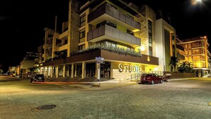 Un immeuble à plusieurs étages, sur lequel on peut voir une grande enseigne indiquant « STUDIO », avec une entrée au niveau du sol, un espace couvert et une voiture rouge garée devant.