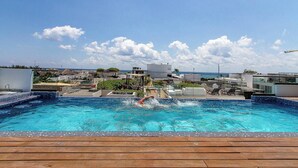 Une piscine où une personne nage, entourée d’une terrasse en bois et avec des bâtiments modernes en arrière-plan.