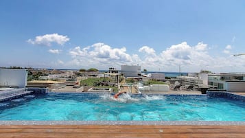 Una piscina con una persona nadando, rodeada de una terraza de madera y edificios modernos al fondo.