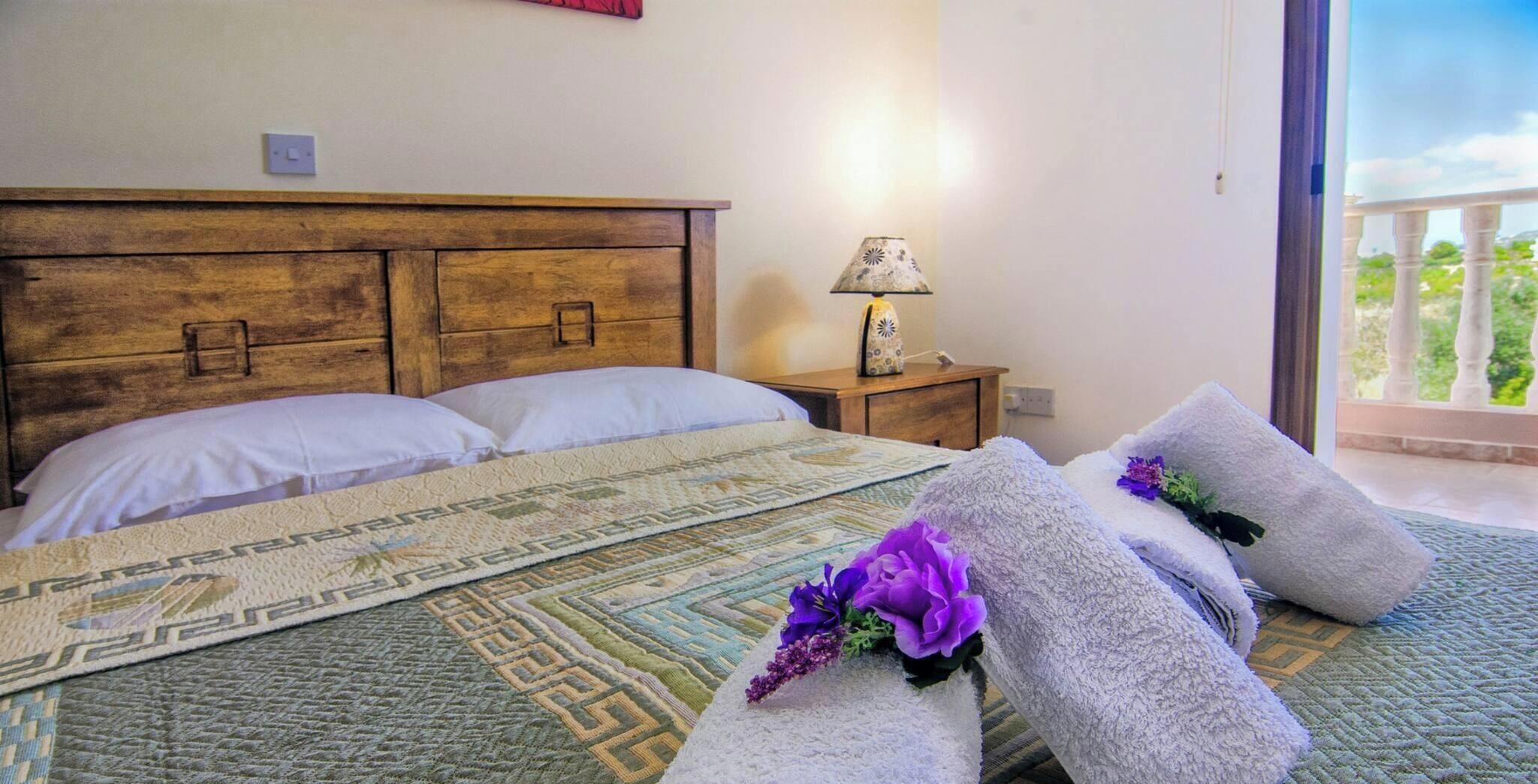 Una camera da letto con una testiera in legno, un letto con lenzuola bianche, una coperta decorata e un comodino con una lampada, oltre a vista su del verde attraverso una finestra.
