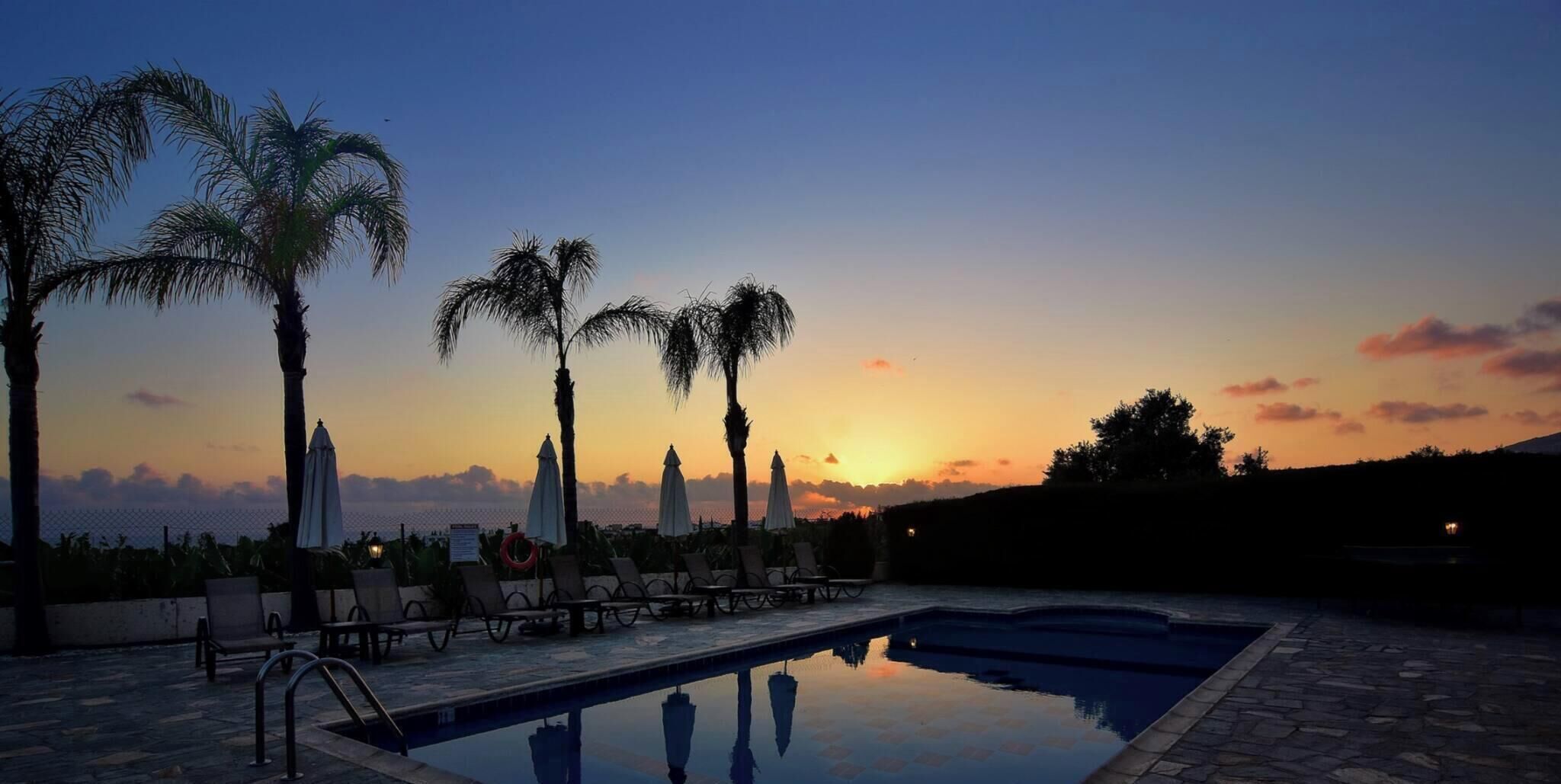 A poolside area with lounge chairs and umbrellas, palm trees, and a sunset view.