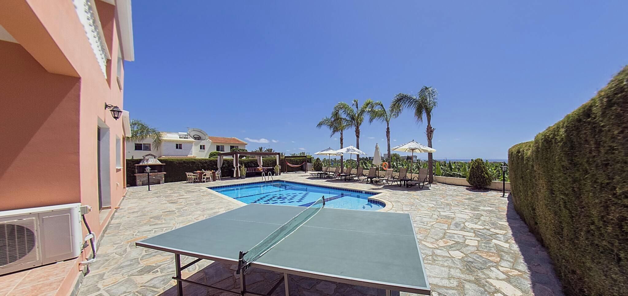 Una piscina con sillas de descanso y sombrillas, una mesa de ping pong y un seto bien cuidado.