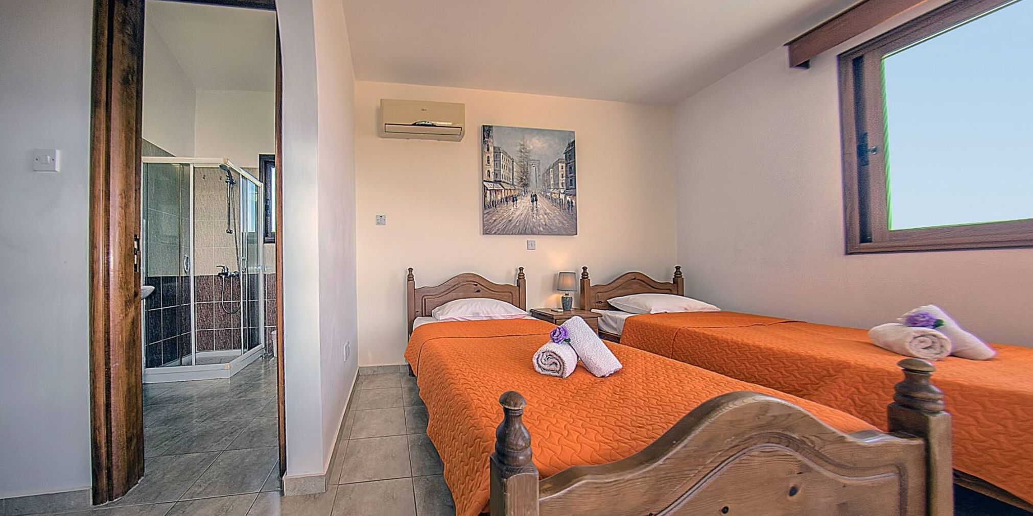 A hotel room with two beds, a shower, a painting on the wall, and a window.
