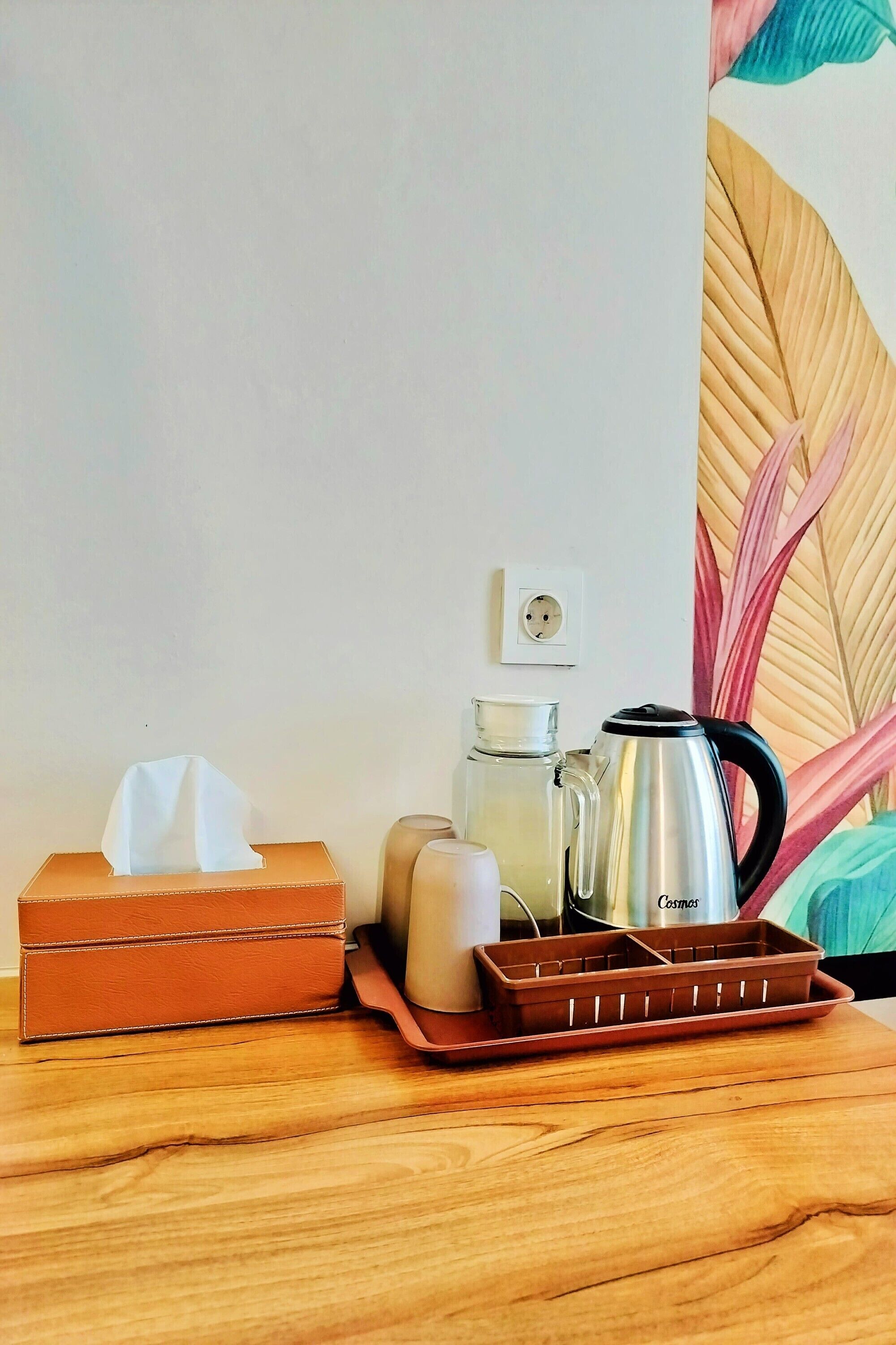 Una mesa de madera con una caja de pañuelos, una jarra de vidrio, una taza y una cafetera de acero inoxidable.