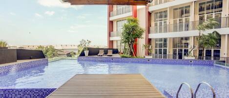 A rooftop swimming pool with wooden deck, lounge chairs, and a modern building with large windows in the background.