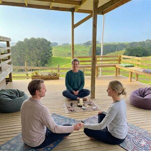 A wooden deck with a view of green fields, a wooden pergola, and a person sitting on a mat. - Tranquility and comfort. (Awhitu)