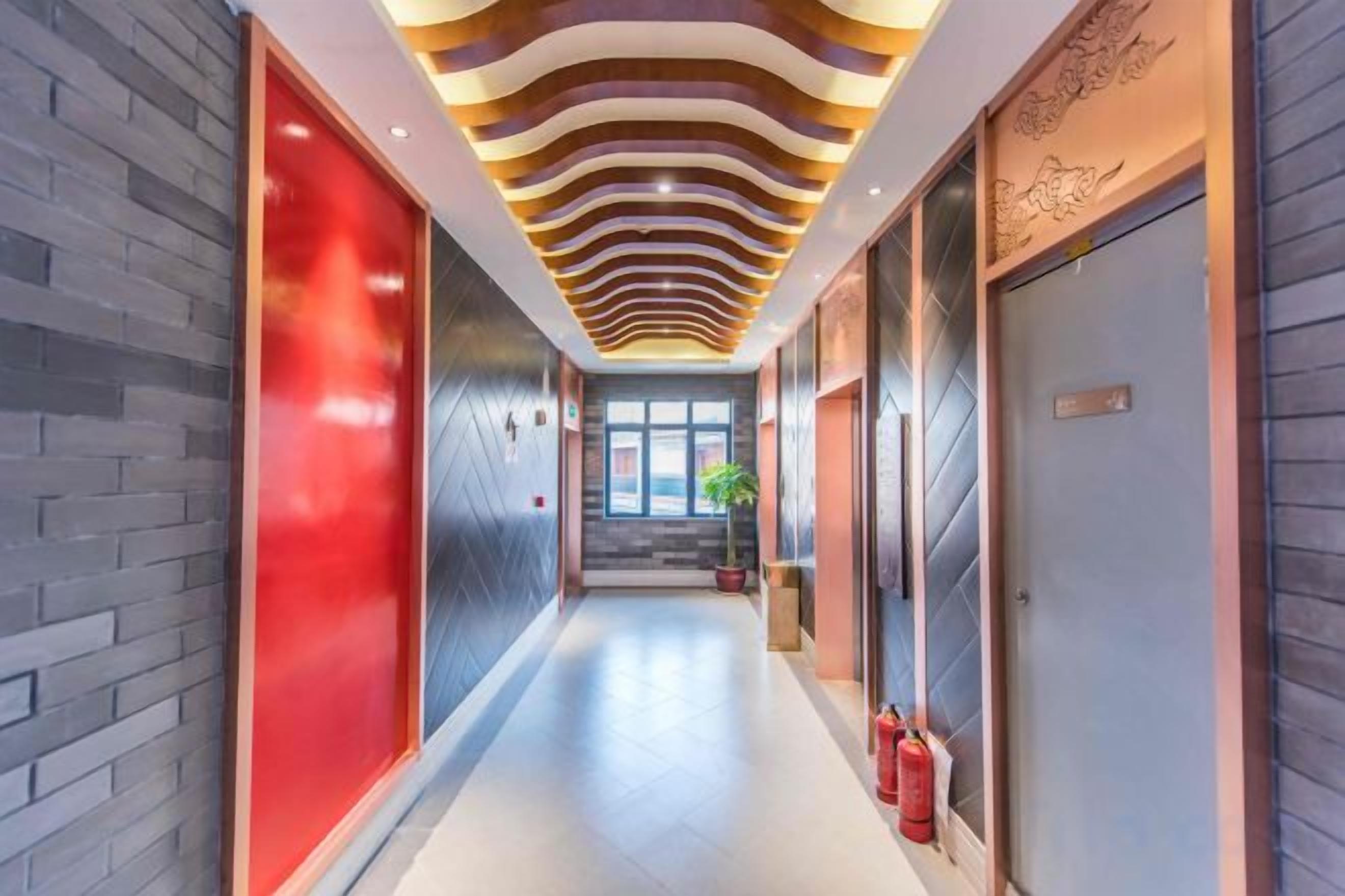 A hotel corridor with a modern design, featuring a patterned ceiling, a fire extinguisher, and a potted plant.
