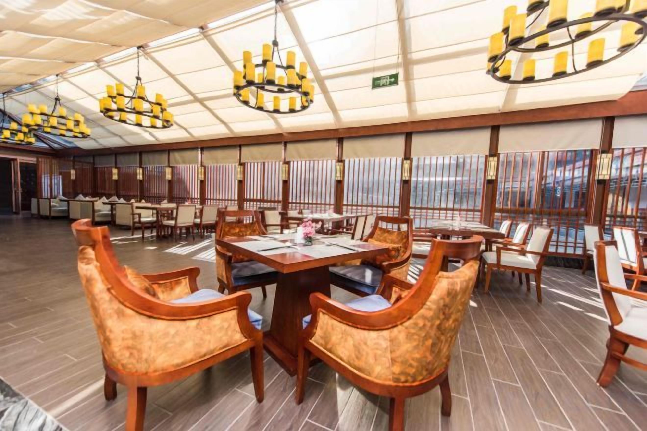 A spacious dining area with multiple tables and chairs, a high ceiling with chandeliers, and large windows.