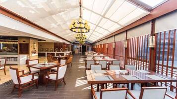 A spacious restaurant with high ceilings and a glass roof, featuring wooden tables and chairs, and a central chandelier.