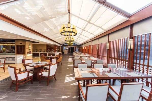 Un restaurante espacioso con techos altos y un techo de cristal, con mesas y sillas de madera y una lámpara central.