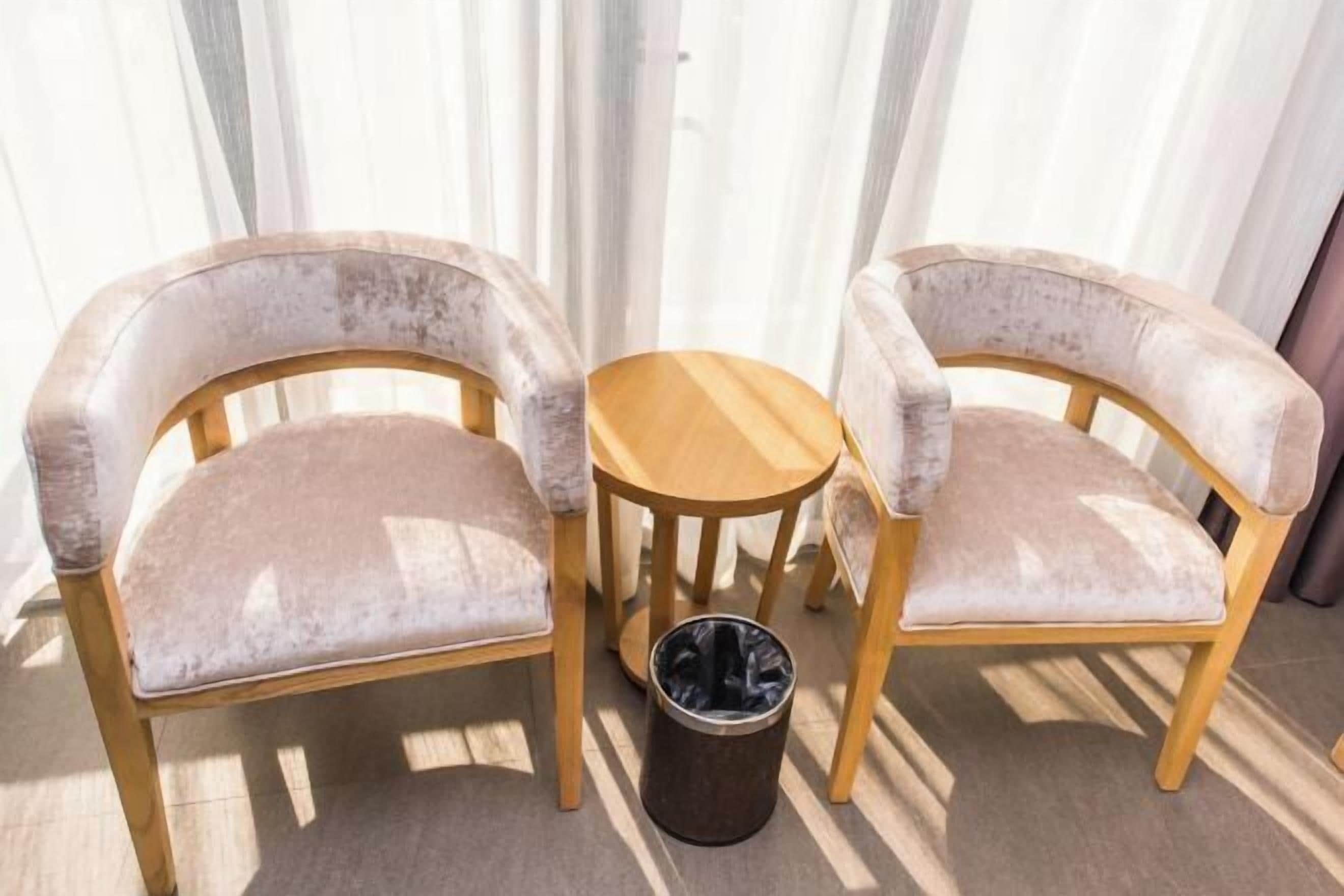Dos sillones tapizados con estructura de madera y una pequeña mesa de madera, sobre un piso de baldosas, con un cesto de basura cerca.