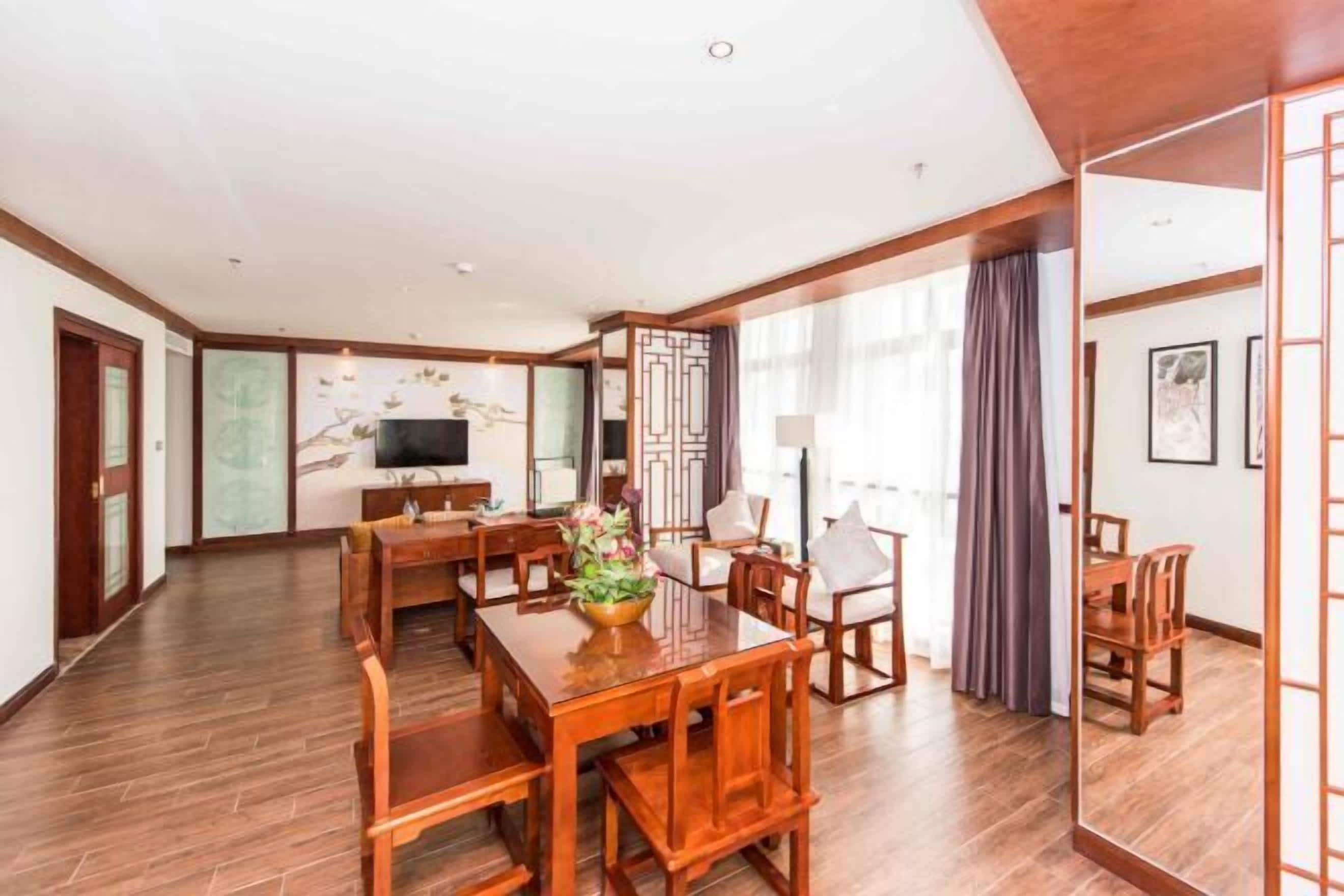 A spacious hotel room with a wooden dining table, chairs, a TV, and a large window with curtains.